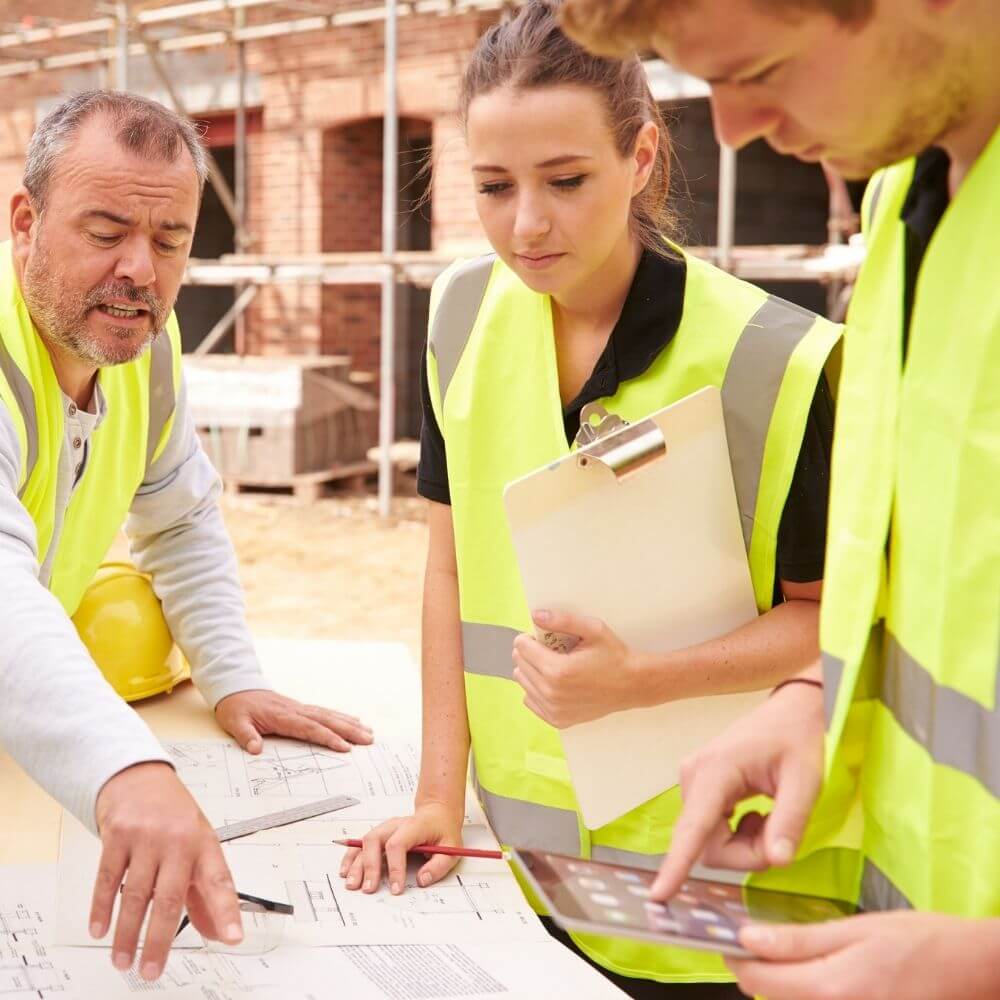 Füllbild: Zwei Männer und eine Frau lehnen sich auf einer Baustelle über Pläne.